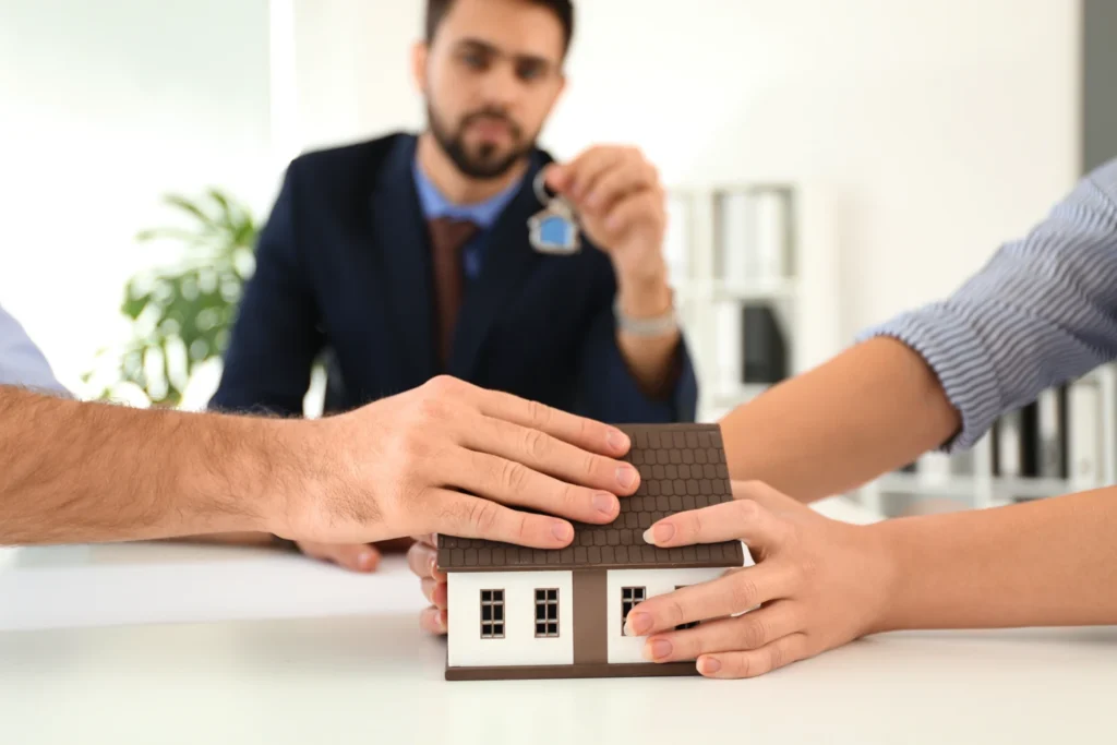 A Couple Reaching for a model house representing their community property, with a realtor in between, handing them the keys to the house